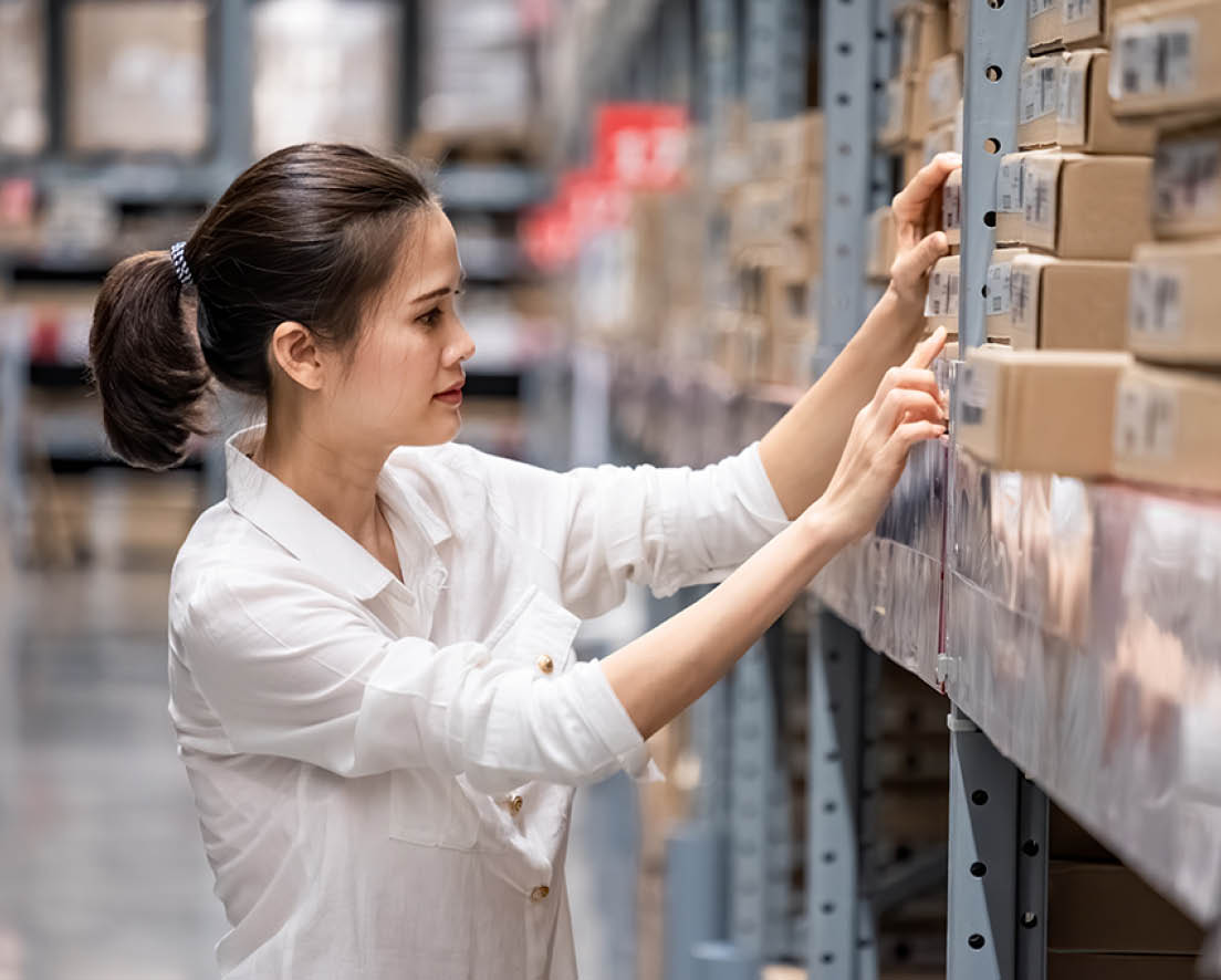 Smart Asian young woman happy to work in store warehouse. Girl standing and checking quality and quantity of inventory stock on shelf by model name and serial number it show on packaging of goods.